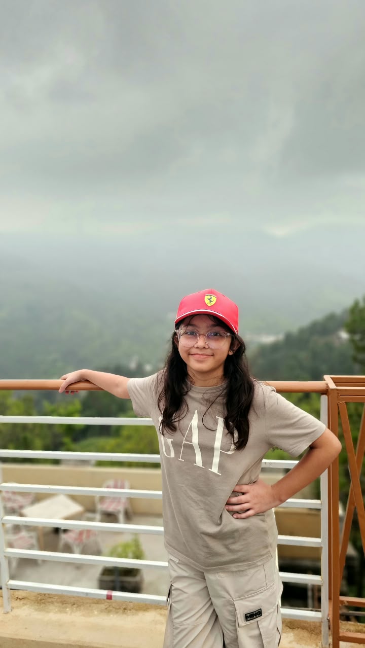 Enayah smiling in a red Ferrari cap on a mountain-view balcony